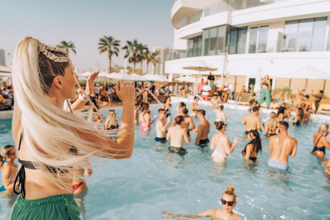 Five Jumeirah Village Dubai Exterior foto The photo features a lively pool party scene. In the foreground, a woman with long, blonde hair is seen raising her arm as if enjoying the moment. The pool is filled with many people, some swimming and others socializing. The atmosphere appears festi