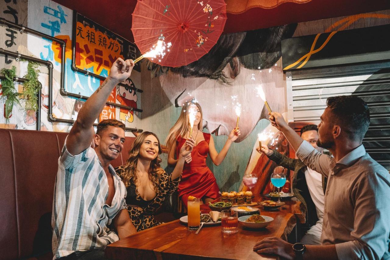 Five Jumeirah Village Dubai Exterior foto The photo depicts a lively gathering at a dining table where several people are celebrating. There are four individuals: two men and two women. One of the women is wearing a red dress, while the other is dressed in a patterned outfit. They are holdin