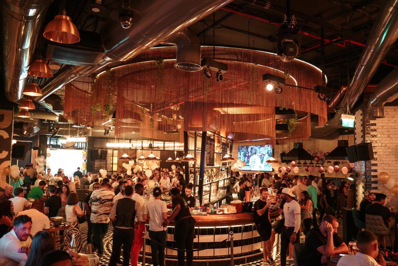 Five Jumeirah Village Dubai Exterior foto The photo depicts a lively bar or restaurant scene filled with people socializing. The interior design features a modern aesthetic with a mix of industrial elements, such as exposed pipes and metallic accents. A large, circular bar is the focal point