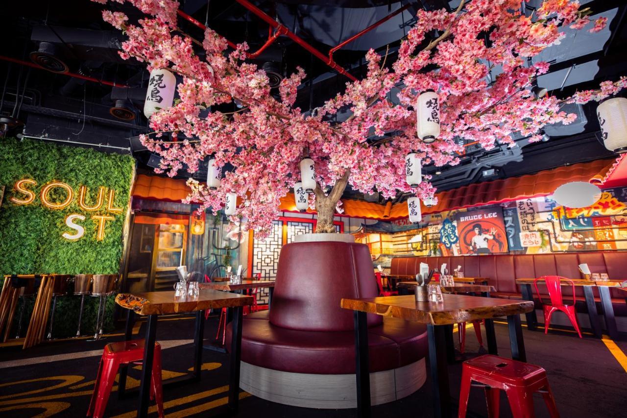 Five Jumeirah Village Dubai Exterior foto The photo shows a vibrant and modern restaurant interior. At the center, there is a large cherry blossom tree adorned with pink flowers and hanging lanterns, creating a lively atmosphere. Surrounding the tree is a circular booth with maroon upholster