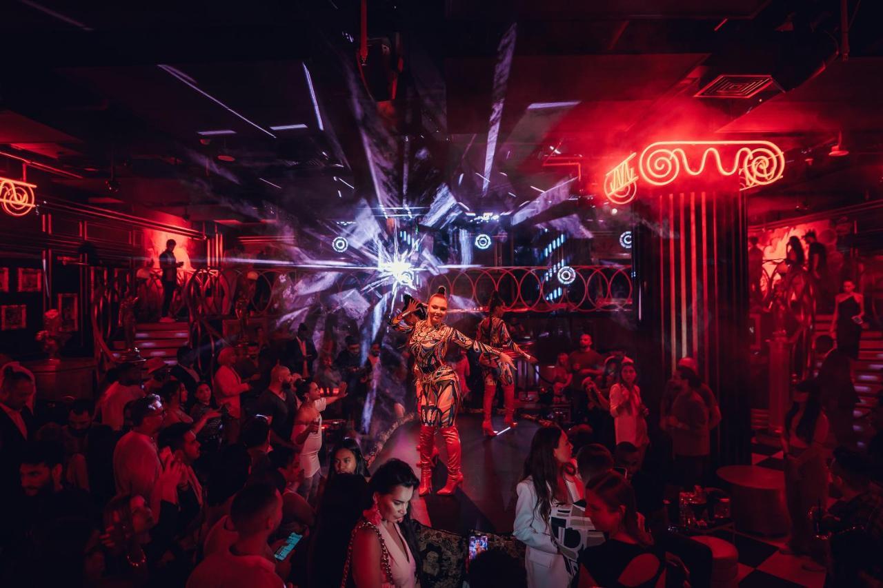 Five Jumeirah Village Dubai Exterior foto The photo depicts a vibrant nightlife scene, likely from a nightclub or performance venue. The setting is dimly lit with striking red lights and dynamic visual effects, creating an electrifying atmosphere. In the center, a performer dressed in a flas