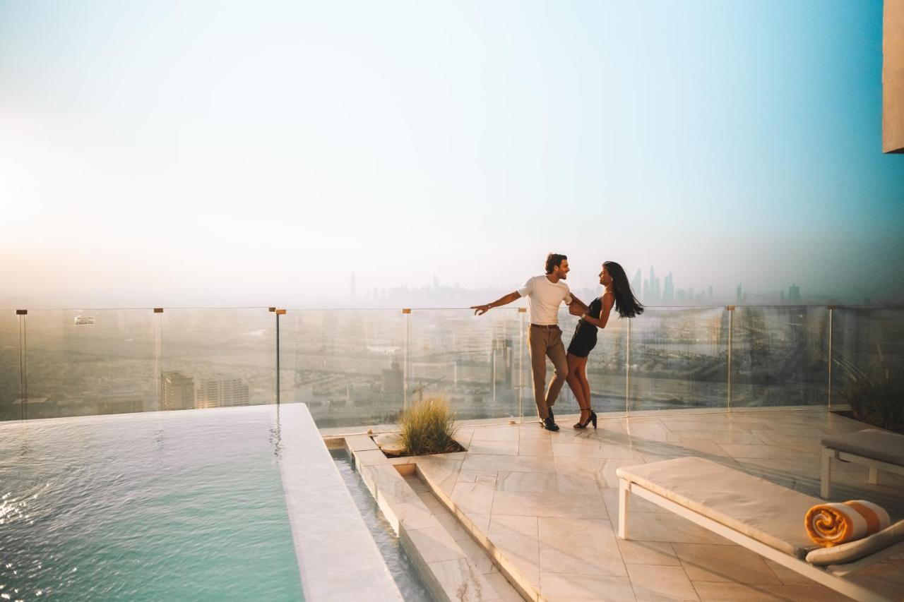Five Jumeirah Village Dubai Exterior foto The image shows a couple dancing together on a terrace or balcony with a view of a city skyline in the background. They appear to be enjoying a romantic moment, surrounded by a luxurious setting that includes a swimming pool. The atmosphere is bright