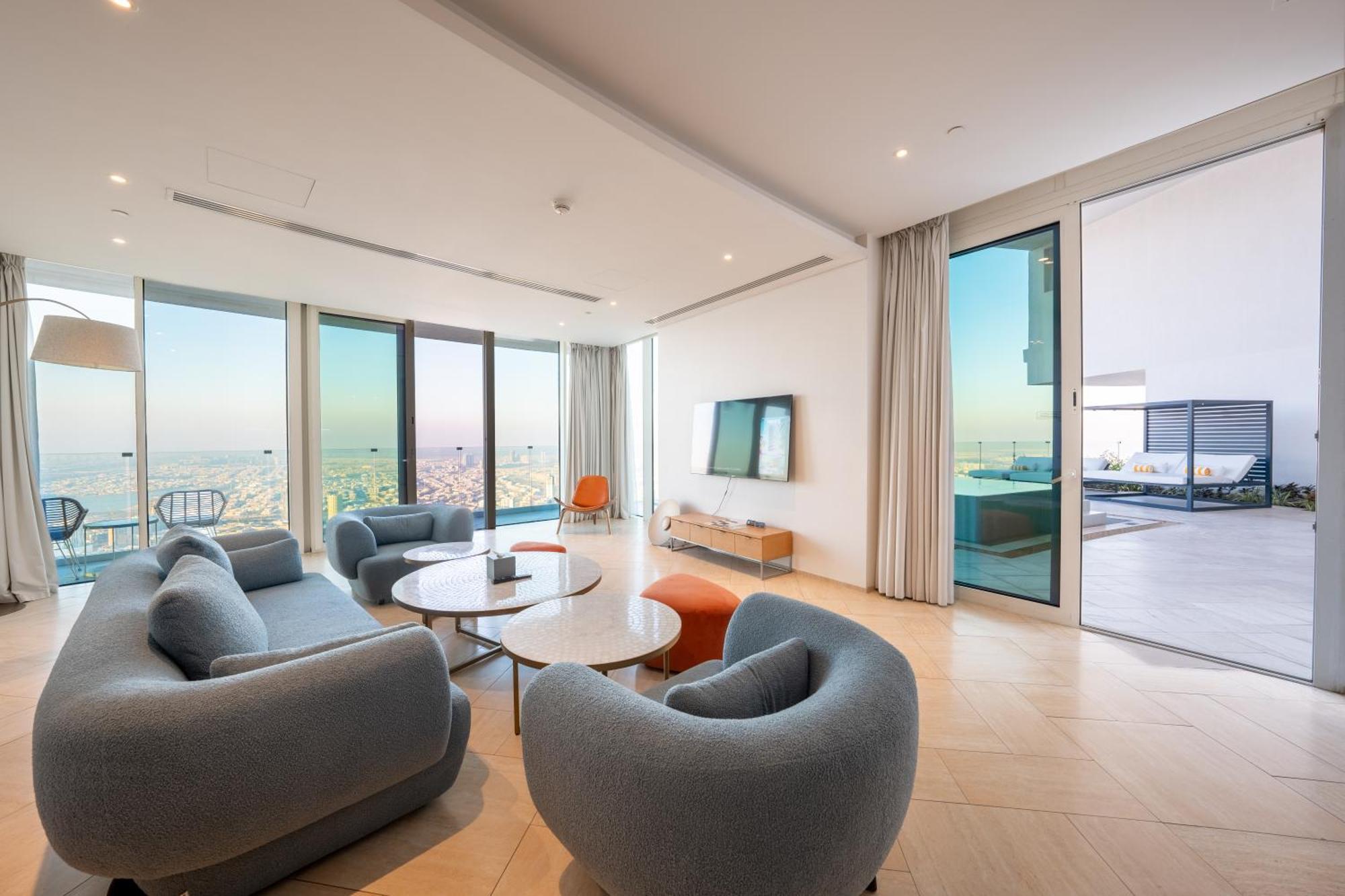 Five Jumeirah Village Dubai Exterior foto The photo shows a modern and stylish living room area with large windows that provide a scenic view. There are several rounded, gray sofas arranged around a couple of round coffee tables. A sleek television is mounted on the wall across from the seat