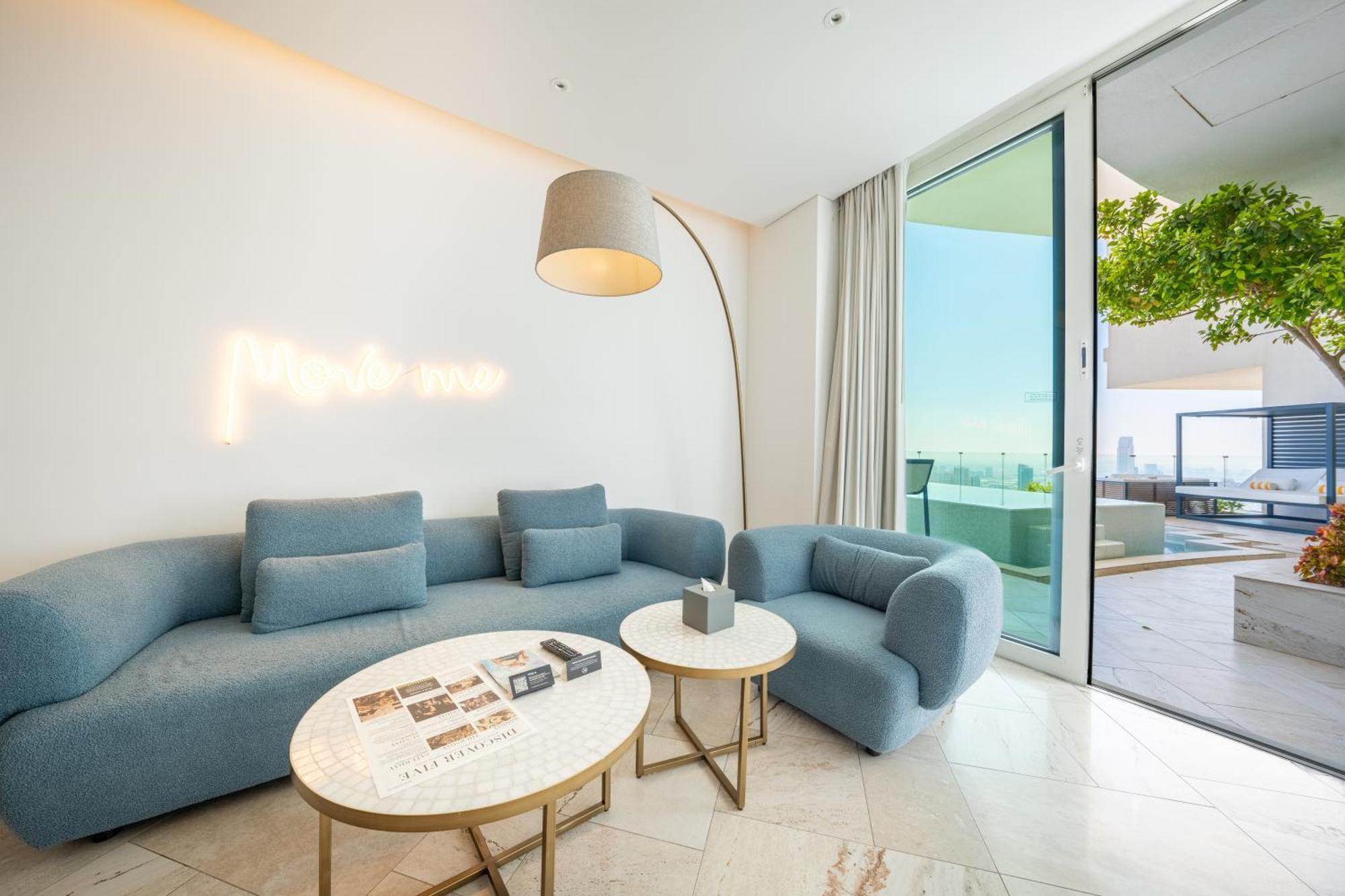 Five Jumeirah Village Dubai Exterior foto The photo shows a modern and stylish living space. It features a light blue sectional sofa and a matching armchair, creating a cozy seating area. Two round marble coffee tables are placed in front, one of which has some magazines or books on it. A ta