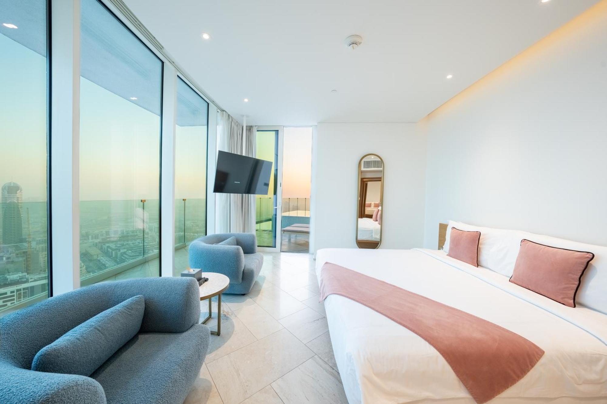 Five Jumeirah Village Dubai Exterior foto The photo depicts a modern hotel room featuring large windows that provide a view of the outside. The room includes a spacious bed with a pink accent blanket, a cozy blue sofa, a small round table, and a wall-mounted television. There is also a mirro