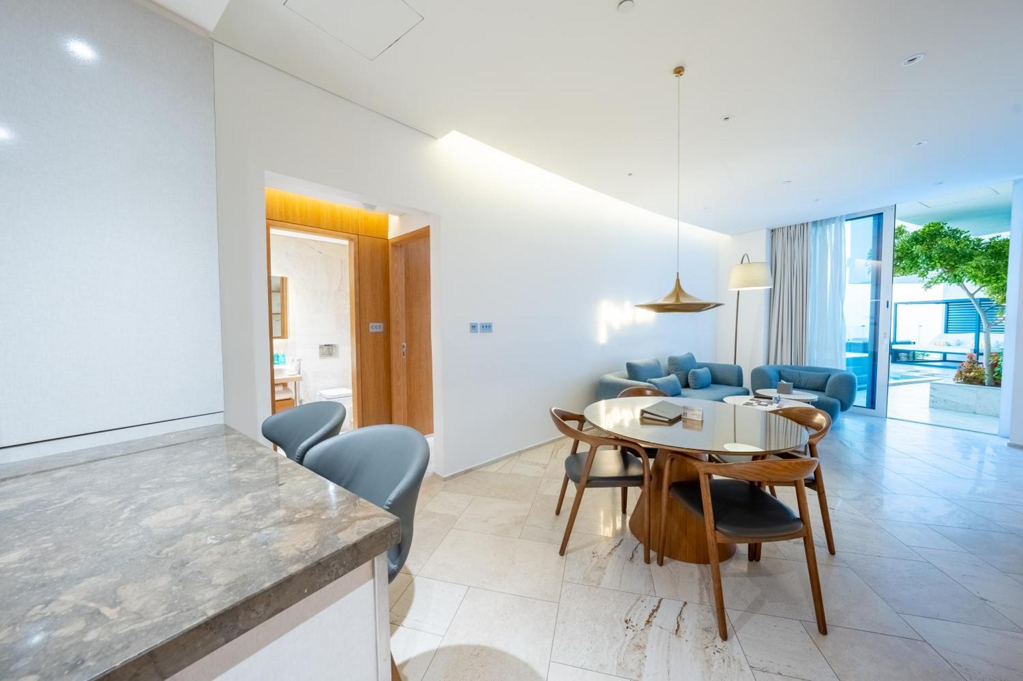 Five Jumeirah Village Dubai Exterior foto The photo shows a modern living space that combines a dining area and a lounging area. On the left, there is a kitchen counter with a marble surface and several gray chairs around it. In the center, there is a round dining table made of wood with sev