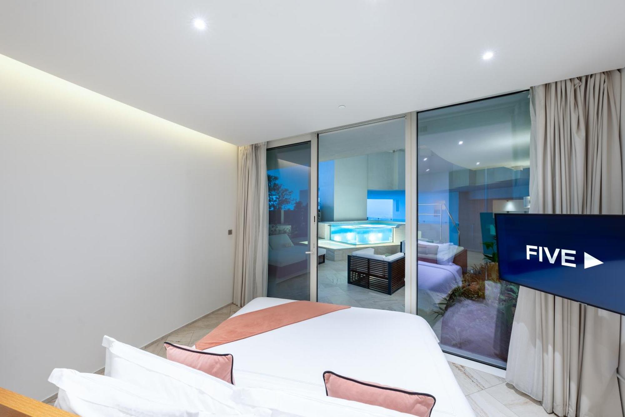 Five Jumeirah Village Dubai Exterior foto The photo shows a modern, stylish bedroom featuring a large bed with light-colored bedding and decorative pillows. There are large glass windows that open up to a view of an outdoor area, likely overlooking a pool or patio. The room has neutral-color