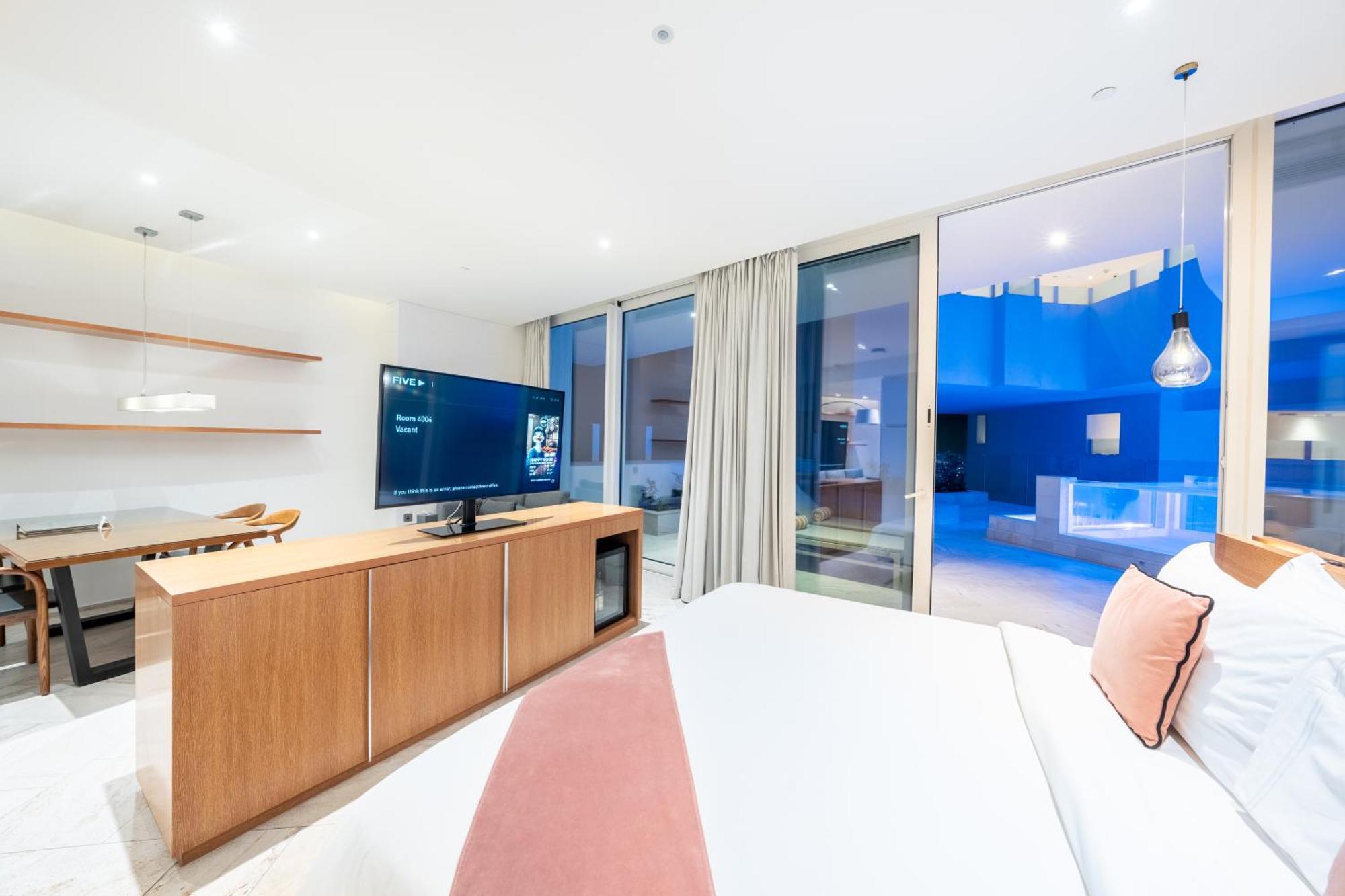 Five Jumeirah Village Dubai Exterior foto The photo depicts a modern hotel room with a clean and minimalist design. A large bed is shown, topped with white linens and decorative pillows. Opposite the bed, there is a wooden TV stand with a flat-screen television. The room features large windo