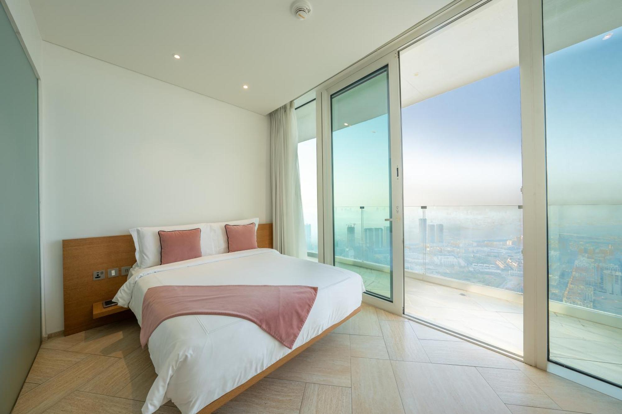 Five Jumeirah Village Dubai Exterior foto The photo shows a modern bedroom with a large bed featuring white bedding and accent pillows in a soft pink color. The room has floor-to-ceiling glass doors that lead out to a balcony, providing a panoramic view of the outside. The walls are painted 