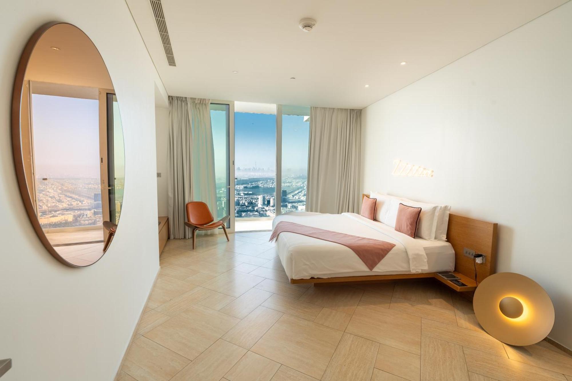 Five Jumeirah Village Dubai Exterior foto The photo shows a modern bedroom with a minimalist design. There is a large bed with white bedding and pink accents, positioned against a wall. To the side, there is a round mirror and a stylish chair. The room features large windows dressed with lig
