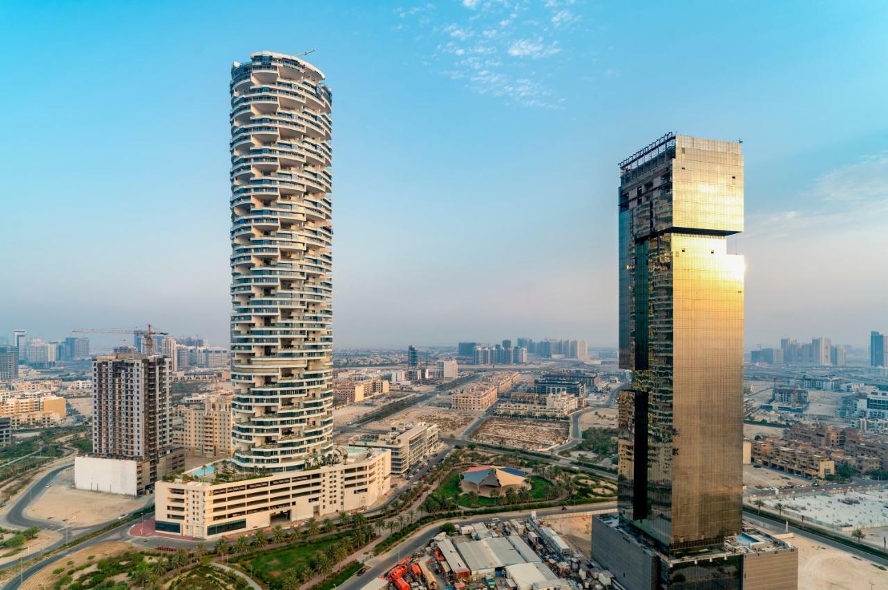 Five Jumeirah Village Dubai Exterior foto The photo shows two distinctive skyscrapers located in an urban area. On the left, there is a tall, cylindrical building with a unique, modern design characterized by its twisting, spiral shape and balconies at various levels. To the right, there is 