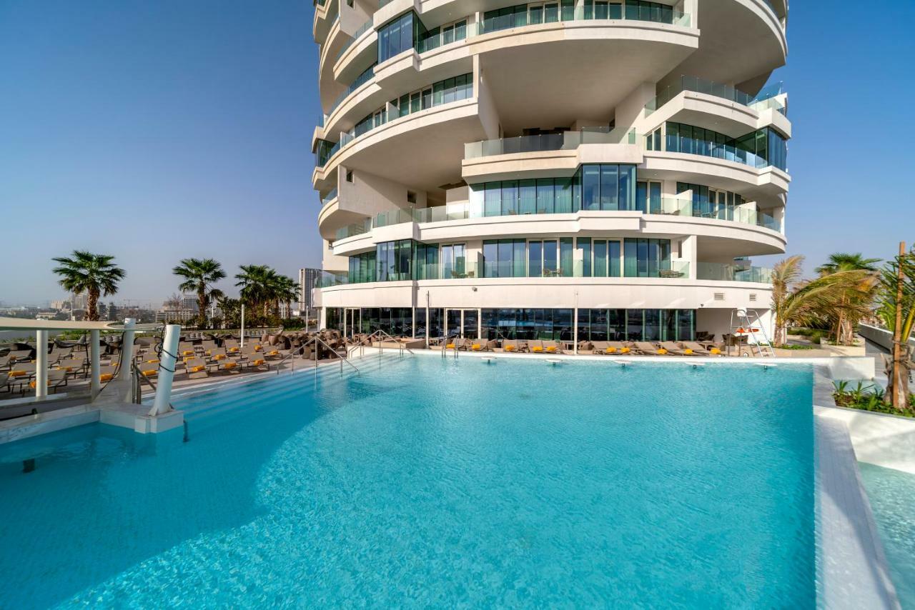 Five Jumeirah Village Dubai Exterior foto The photo depicts a modern building with a distinctive architectural design, featuring multiple curved balconies. In front of the building, there is a large, inviting swimming pool with clear, turquoise water. Surrounding the pool are loungers for re