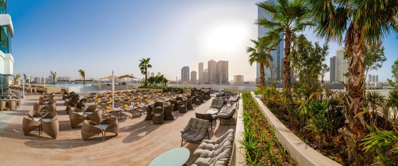 Five Jumeirah Village Dubai Exterior foto The photo showcases a luxurious outdoor seating area, likely part of a resort or hotel. In the foreground, there are well-arranged lounge chairs and seating options, some with cushions, placed on a tiled surface. The area is bordered by lush green pl